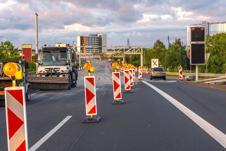 Straße mit Baustellenabsperrungen und Warnleuchten, Lkw und Pkw fahren auf der Fahrbahn, im Hintergrund Gebäude und bewölkter Himmel.