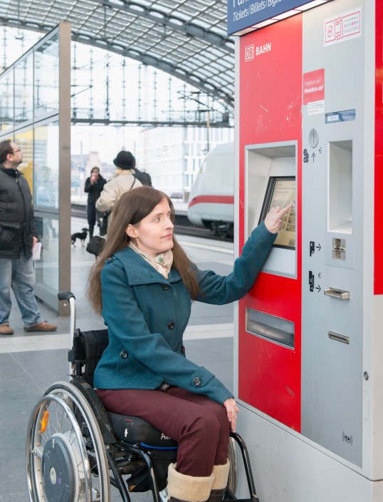 Frau im Rollstuhl bedient Fahrkartenautomat