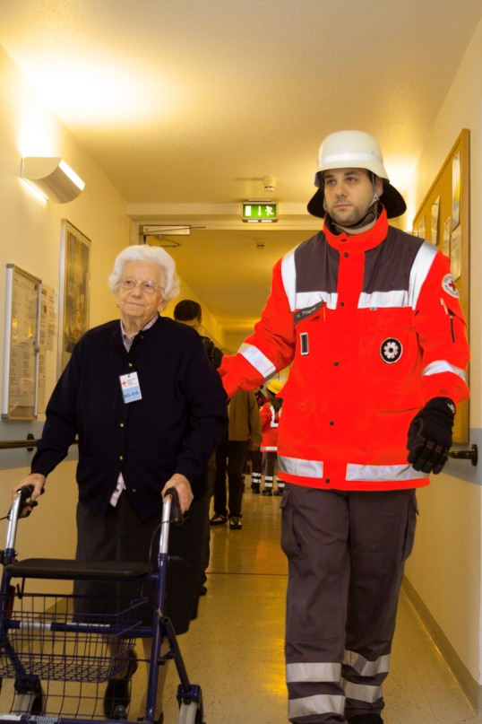 Eine alte Frau mit Rolator wird von einem Rettungssanitäter in Uniform über einen sehr breiten Flur eines Seniorenheims geführt.