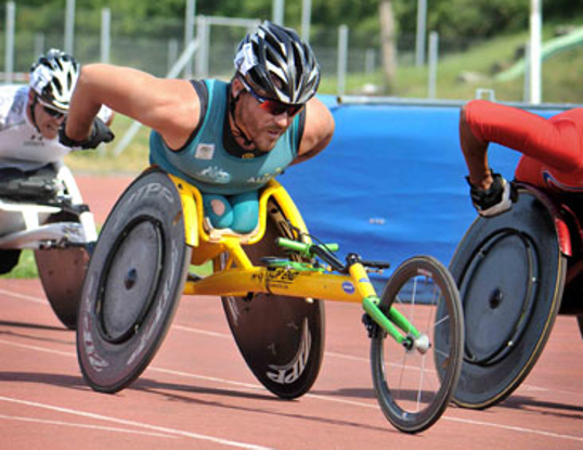 Bild zeigt Kurt Fearnley, der Beinstummel hat in einem Rennrollstuhl, der zwei hinten Räder hat. An einer langen Stange ist vorne ein drittes Rad. Kurt stößt sich mit beiden Armen ab, den Lenker fasst er nicht an.