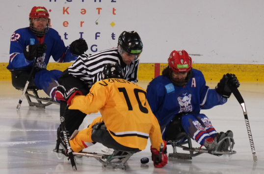 Das Bild zeigt drei Para-Eishockeyspieler und einen Schiedsrichter. Die Spieler sitzen auf einer Art Schlitten. Mit dem Schläger stoßen sie sich auf dem Eis ab.