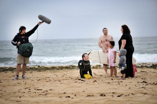 Links steht Toningeneur mit hochgehaltenem Mikro bildmittig Kurt fearnley im Gespräch mit Familie rechts von ihm. Er ist ohne seine Beine kleiner als auch die kleinen Kinder der Familie.