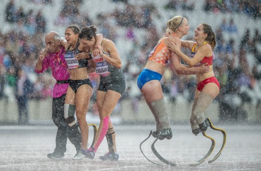 Foto zeigt zwei im Regen feiernde Läuferinnen, während eine dritte enttäuscht, gestützt von zwei Personen den Platz verlässt