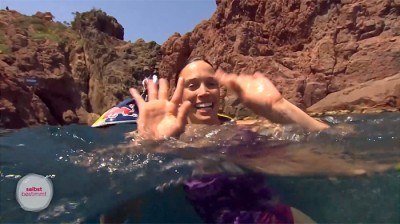 Springerin aufgetaucht, winkt, kahle Felsen im Hintergrund, Sonnenschein, Motorboot etwas versteckt. hinter der Springerin. Kamera halb unter Wasser.