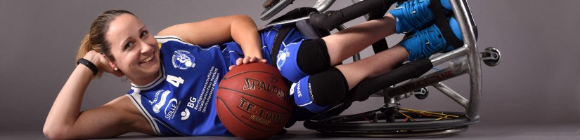 Bild zeigt Frau im umgekippten Rollstuhl sitzend, mit Basketball. Studiobild.