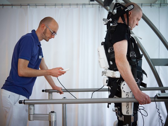 Mann übt am Geländer gehen im Exoskelett, dahinter programmiert ein Physiotherapeut die Gangparameter. Reha-Fachkraft. BG Kliniken Bergmannstrost Halle.