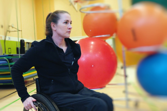 Edina Müller von der Seite im Rollstuhl sitzen. Sie ist im BG Klinikum, Im Hintergrund sind verschiedene Gymnastikbälle zu sehen.