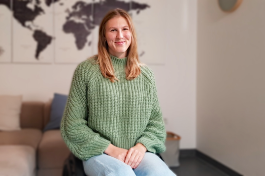 Lilly Sellak zuhause auf einem Stuh sitzend, Wollpullover, Jeans, lächelnd.