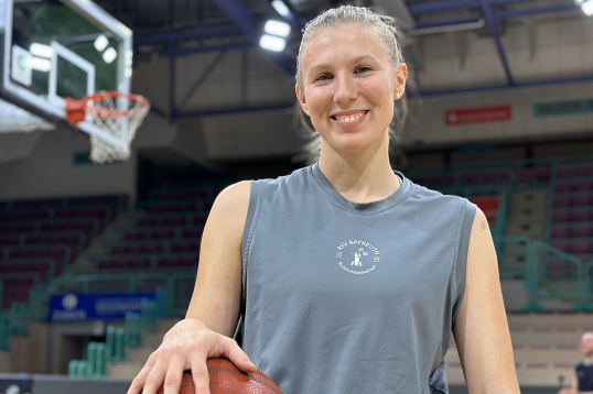 Lilly Sellak im Profil in einer Sporthalle. In der rechten Hand hält sie einen Basketball. Oben links ist verschwommen ein Basketball-Korb zu sehen.