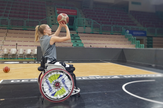 Lilly Sellak in einer Sporthalle im Rollstuhl sitzend, zielt mit den Ball auf dem Korb, der rechts außerhalb des Bildes ist.