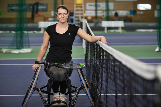 Bild zeigt Britta Wend im Rollstuhl am Netz sitzen. Tennisschläger auf dem Schoß, linker Arm über dem Netz.