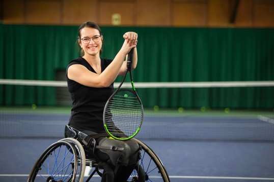 Bild zeigt Britta Wend lächelnd im Rollstuhl vor dem Tennisnetz, den Schläger aufrecht im Schoß.