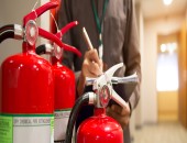 Bild vom oberen Teil von drei Feuerlöschern. Im Hintergrund steht eine Person mit Klemmbrett.