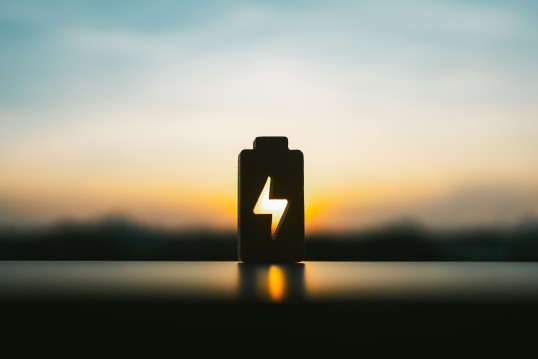 Bild eines Sonnenaufgangs, im Vordergrund ist die Silhouette einer Batterie mit einem Energiesymbol 