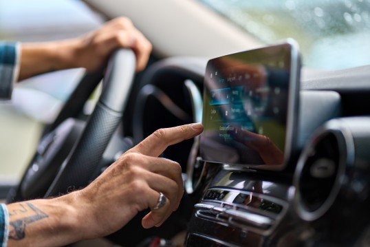 Foto in einem Fahrzeug, man sieht eine Hand die gerade auf einem befestigten Tablet ein Navigationssystem bedient.