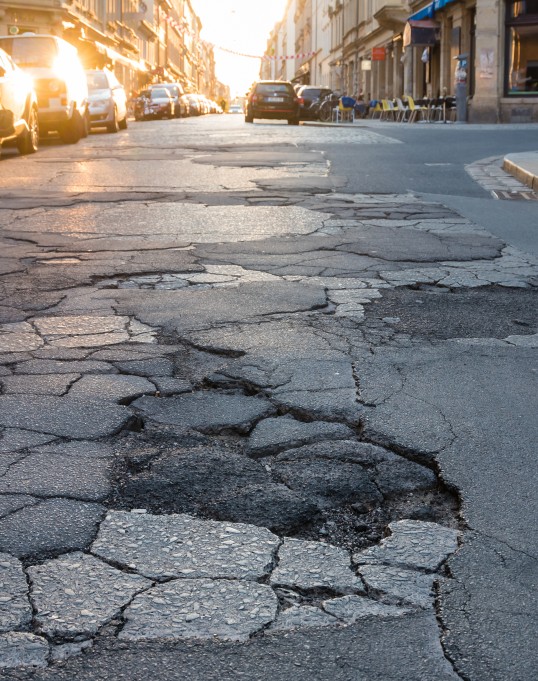 Picture of a road full of potholes, in the background there is a car driving away