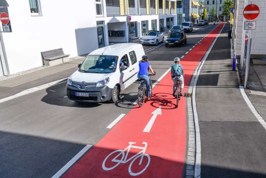 Picture of a Van driving out of a one way street. Two people on bicycles are crossing over the bicycle path which is marked red.