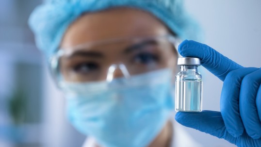 A Picture of a Person wearing latex gloves, a medical mask, a surgical cap and protective glasses. The Person is holding a medical ampoule.