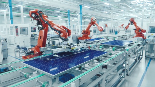 Orangene Industrieroboterarme die in einer modernen und hellen Fabrik an einem Fließband Solarplatten herstellen.