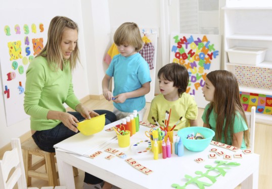 Am Tisch sitzende Erzieherin mit Kindern und Spielgerät