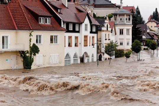 Foto: Überflutete Sraße mit Häuserzeile, gegen die die Wasserfluten drücken.