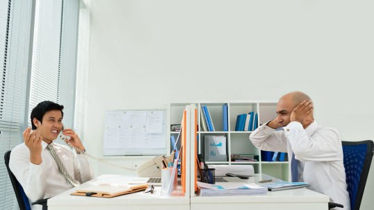 Ein Mann im Büro telefoniert und lacht, der Kollege hält sich die Ohren zu