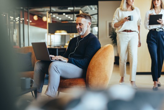 Foto: Mann sitzt in einem Sessel in einer Lobby, auf dem Kopf ein Headset, auf den Knien ein Laptop. Zwei Frauen gehen im Hintergrund vorbei.