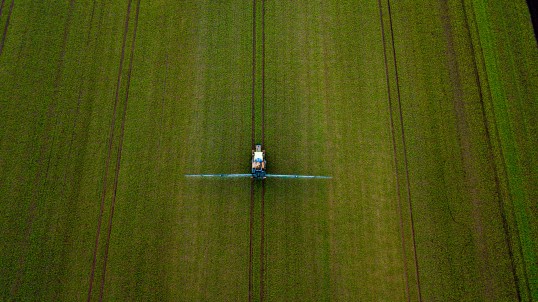 Foto: Feld aus der Vogelperspektive, auf dem ein Traktor mit zwei langen Sprüharmen rechts und links fährt.