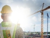 Bild einer Person auf einer Baustelle, die Person trägt einen gelben Bausrellenschutzhelm, im Hintergrund steht ein Kran.