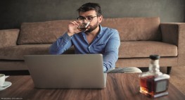 Eine Mann trink ein Glas Alkohol an seinem Homeoffice Arbeitsplatz.