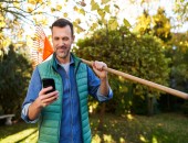 Bild einer Person, die im Garten steht, auf der Schulter eine Harke, in der Hand ein Smartphone.
