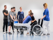 Bild von einer Person, die von einer Pflegefachkraft aus einem Rollstuhl gehoben wird. Eine Person sichert den Rollstuhl, weitere Personen stehen im Hintergrund.