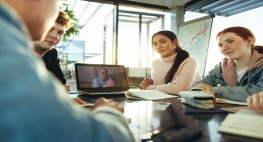 Bild von drei Personen, wie gemeinsam um einen Laptop sitzen, auf dem eine weitere Person zu sehen ist.