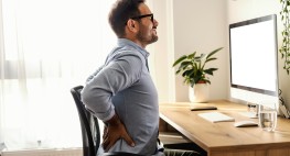 Person sitzt auf einem Schreibtischstuhl vor dem Schreibtisch und fasst mit den Händen in die Seite.