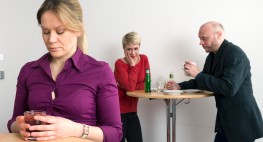 Personen stehen in einem Büro. Zwei Personen sprechen miteinander. Eine Person steht im Vordergrund und schaut nach unten.