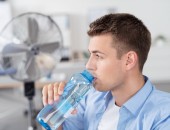 Mann trinkt Wasser im Büro mit Ventilator im Hintergrund.