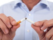 Eine rechte und eine linke Hand zerbrechen eine Zigarette, ein Teil des Oberkörpers einer Person mit einem blauen Hemd ist im Hintergrund zu sehen.