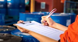 Eine Person füllt eine Checkliste aus, während im Hintergrund Tonnen mit Gefahrstoffen zu sehen sind.