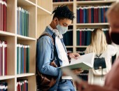 männlicher Student mit Mund-Nase-Bedeckung steht in Bibliothek und leist in Buch