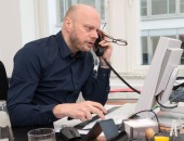ein Mann sitzt am Schreibtisch, er sieht gestresst aus: Telefonhörer am linken Ohr, Blick am Bildschirm und Brille in der linken Hand