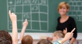 Bild: Schülerinnen und Schüler sitzen an Schulbänken und zeigen auf. Ihnen gegenüber steht eine Lehrerin an einer Tafel.