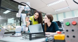 Bild: zwei junge Frauen in einem Techniklabor. Eine Frau sitzt an einem Laptop, die zweite Frau steht in gebeugter Haltung neben ihr und beide haben ihre Blicke auf einen Roboterarm gerichtet, an dem die zweite Frau ein Objekt anbringt.