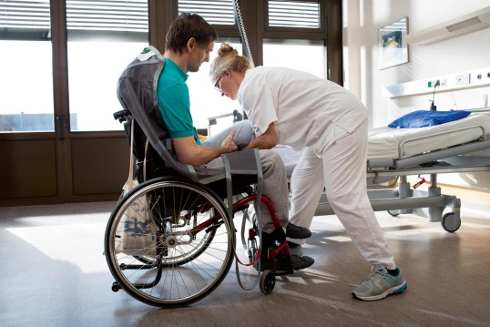 eine Pflegekraft hilft einem Gehbehinderten aus dem Rollstuhl