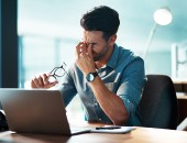 Man sitzt vor einem Computerbildschirm und reibt sich die Augen, dabei hält er eine Brille in der Hand.