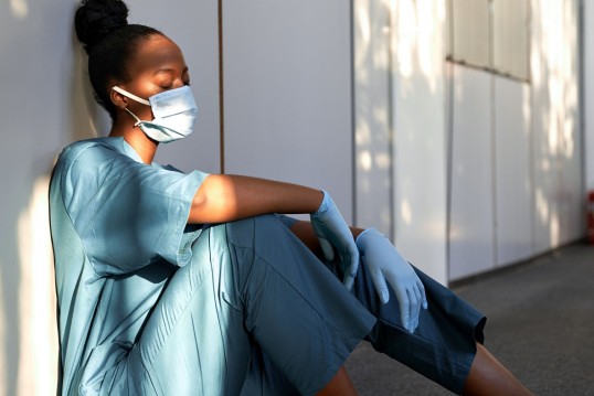 Eine Frau in medizinischer Schutzkleidung sitzt mit geschlossenen Augen an eine Wand angelehnt und wirkt müde.