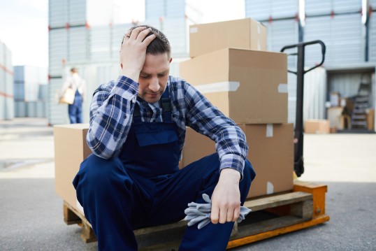 Ein Logistik-Mitarbeiter sitzt auf einem Wagenheber und stützt mit einer Hand seinen Kopf ab.
