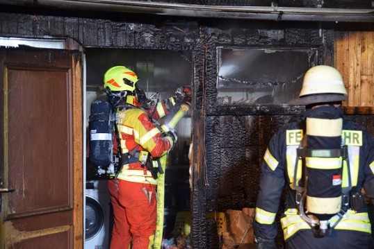Zwei Einsatzkräfte der Feuerwehr löschen einen Hausbrand. Dabei tragen sie ihre funktionsfähige Schutzkleidung sowie umluftunabhängige Atemschutzgeräte.
