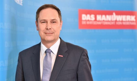 Das Foto zeigt ein Halbporträt von Karl-Sebastian Schulte, Geschäftsführer des ZDH. Er steht im Dunklen Anzug vor einem blauen Aufsteller, auf dem das Logo des Handwerks zu sehen ist.