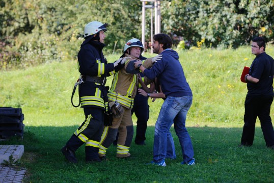 Zu sehen sind mehrere Feuerwehrmänner und Zivilpersonen. Ein Feuerwehrmann hält eine Zivilperson zurück, welche versucht einen Feuerwehrmann anzugreifen.