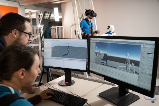 Zu sehen sind zwei Personen vor zwei Bildschirmen, die sich eine Gang-Computersimulation anschauen. Im Hintergrund sieht man die eine Testperson in einem Ganzkörpermessanzug auf einem Laufband.